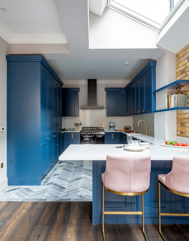 This is an example of a mid-sized transitional u-shaped open plan kitchen in London with a farmhouse sink, shaker cabinets, blue cabinets, marble benchtops, white splashback, marble splashback, black appliances, porcelain floors, a peninsula, blue floor and white benchtop.