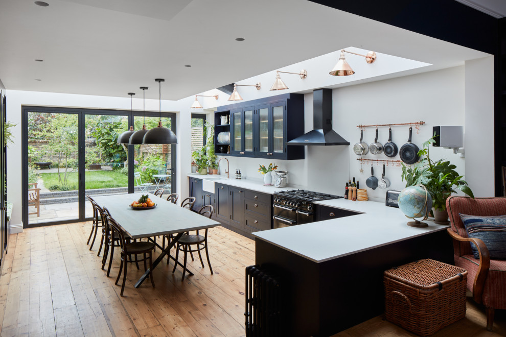Photo of a large eclectic l-shaped kitchen/diner in London with a belfast sink, shaker cabinets, blue cabinets, engineered stone countertops, white splashback, engineered quartz splashback, black appliances, light hardwood flooring, no island, grey worktops, exposed beams and feature lighting.