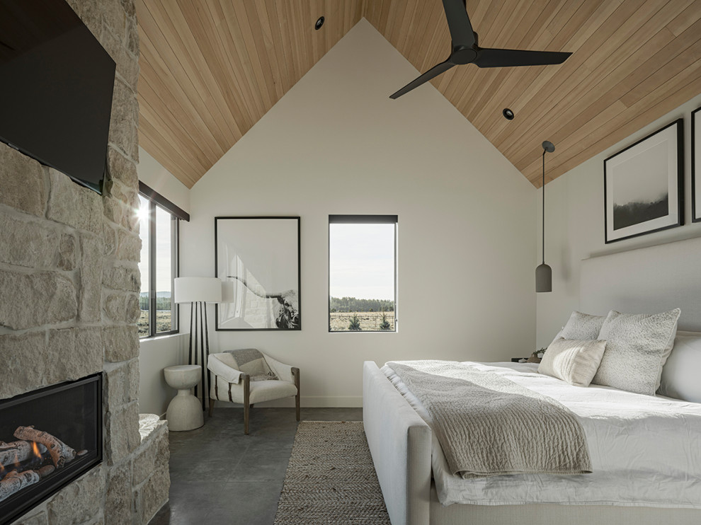 На фото: хозяйская спальня в стиле кантри с белыми стенами, бетонным полом, угловым камином, фасадом камина из камня, серым полом и сводчатым потолком