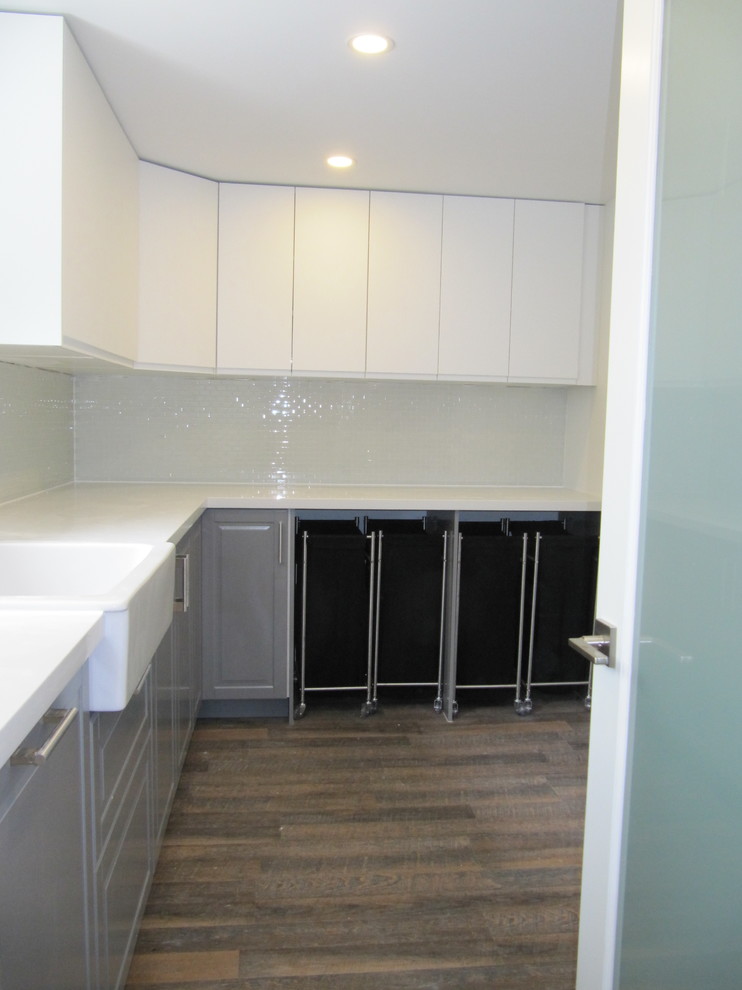Photo of a mid-sized modern l-shaped utility room in Toronto with a farmhouse sink, flat-panel cabinets, grey cabinets, quartzite benchtops, white walls, vinyl floors, a stacked washer and dryer, brown floor and white benchtop.