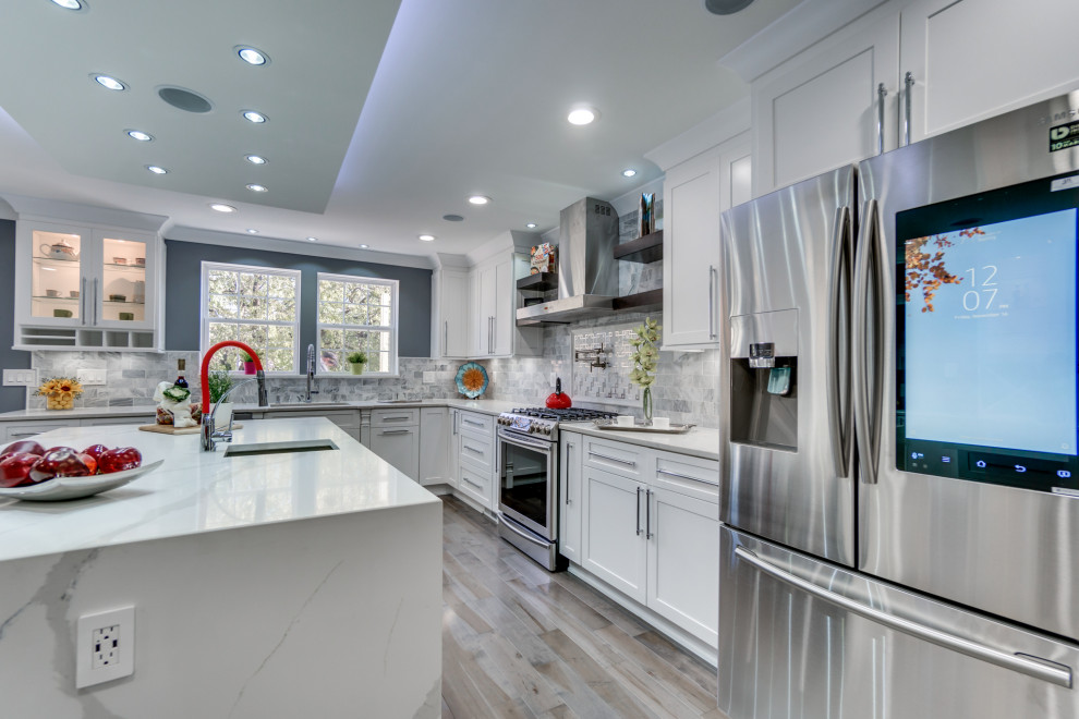 Modern inredning av ett stort vit vitt kök, med en nedsänkt diskho, skåp i shakerstil, vita skåp, marmorbänkskiva, grått stänkskydd, stänkskydd i mosaik, rostfria vitvaror, plywoodgolv, en köksö och grått golv