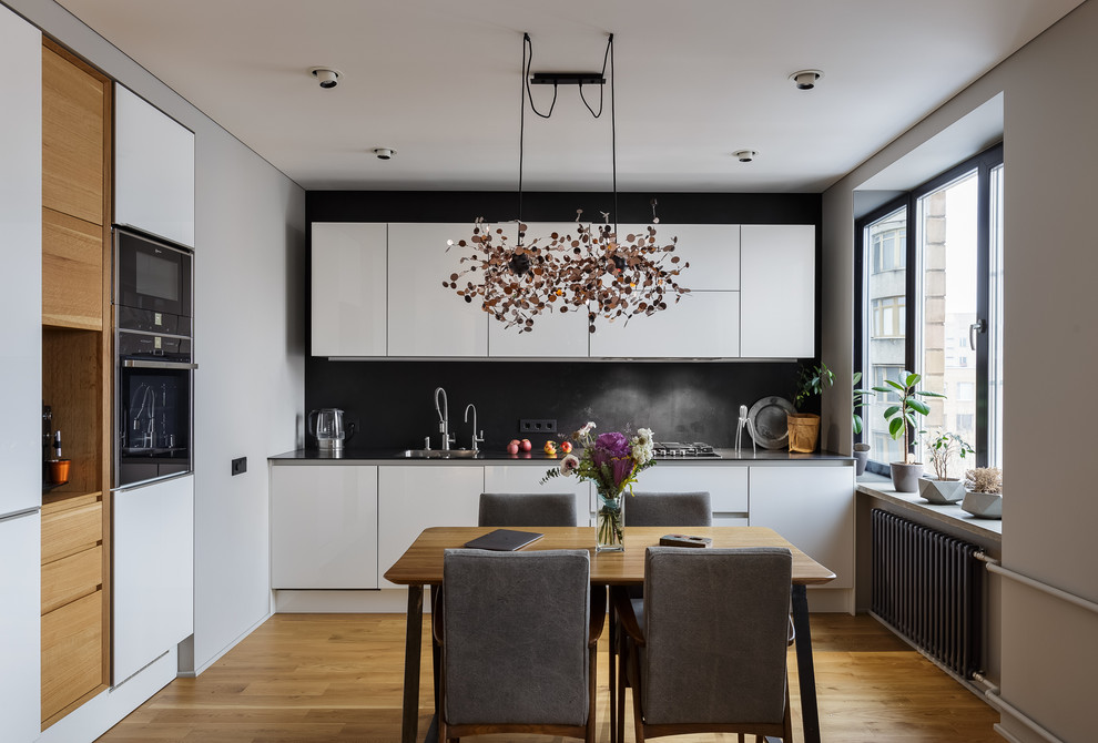 Contemporary eat-in kitchen in Moscow with flat-panel cabinets, white cabinets, black splashback, no island, black appliances, medium hardwood floors and an undermount sink.