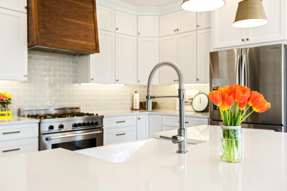 Example of a transitional l-shaped light wood floor and brown floor kitchen design with a farmhouse sink, shaker cabinets, white cabinets, quartz countertops, white backsplash, subway tile backsplash, stainless steel appliances, an island and white countertops