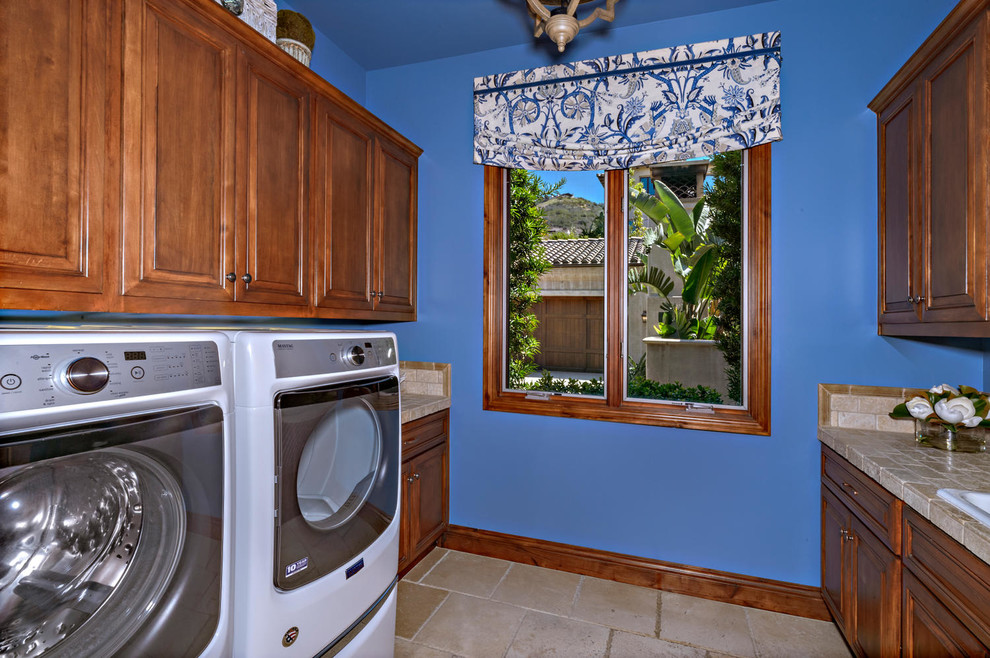 Inspiration for a mid-sized beach style dedicated laundry room in San Diego with a drop-in sink, raised-panel cabinets, brown cabinets, tile benchtops, blue walls, travertine floors, a side-by-side washer and dryer, beige floor and beige benchtop.