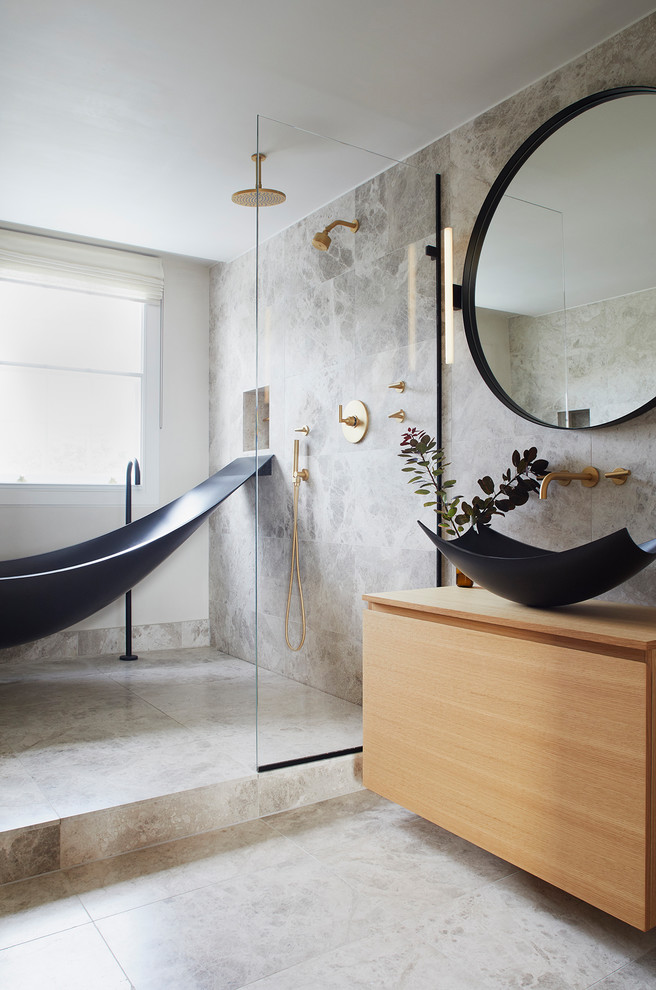 This is an example of a mid-sized contemporary master wet room bathroom in London with light wood cabinets, a freestanding tub, a wall-mount toilet, gray tile, limestone, grey walls, limestone floors, a vessel sink, wood benchtops, grey floor, an open shower, beige benchtops and flat-panel cabinets.