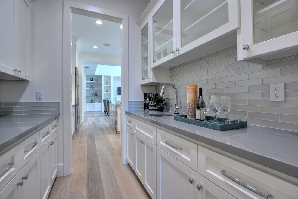 This is an example of a large transitional l-shaped kitchen pantry in San Francisco with a farmhouse sink, shaker cabinets, white cabinets, quartz benchtops, glass tile splashback, stainless steel appliances, light hardwood floors, with island and grey splashback.