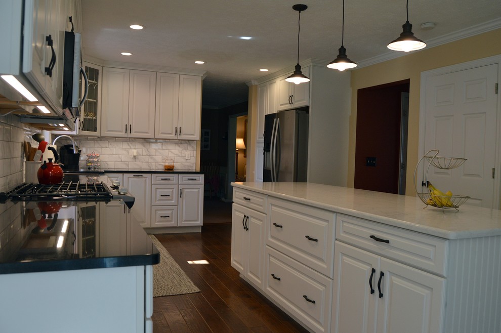 Photo of a mid-sized transitional l-shaped eat-in kitchen in Other with a farmhouse sink, raised-panel cabinets, white cabinets, quartz benchtops, white splashback, stainless steel appliances, medium hardwood floors, with island, brown floor and white benchtop.