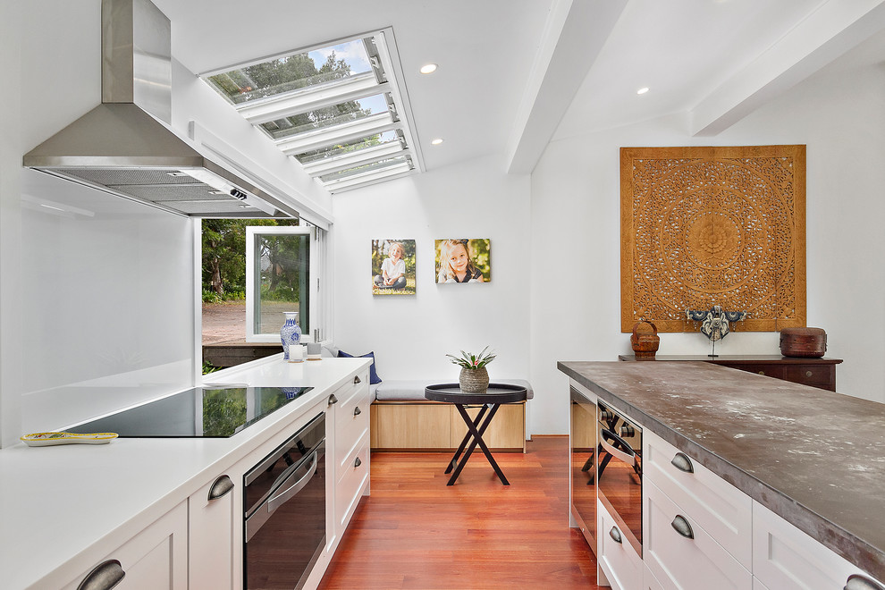 Expansive contemporary l-shaped open plan kitchen in Sydney with an integrated sink, shaker cabinets, white cabinets, zinc benchtops, white splashback, glass sheet splashback, black appliances, light hardwood floors, with island, brown floor and grey benchtop.