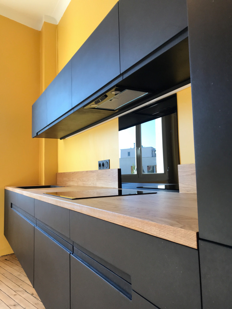Photo of a mid-sized contemporary single-wall open plan kitchen in Bordeaux with an undermount sink, flat-panel cabinets, black cabinets, wood benchtops, beige splashback, timber splashback, stainless steel appliances, light hardwood floors, no island, beige floor and beige benchtop.