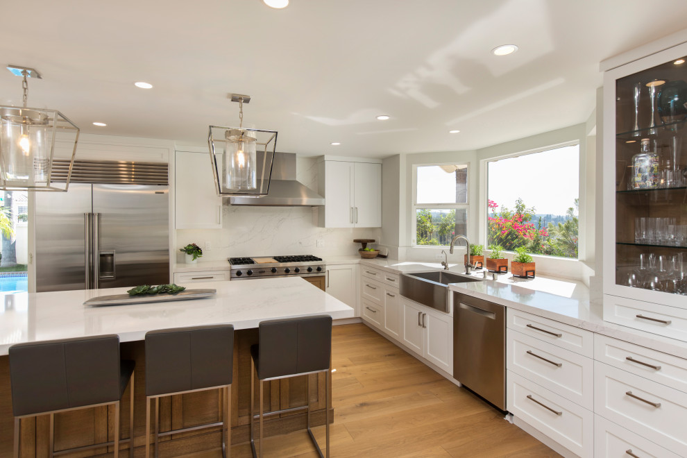 This is an example of a large traditional l-shaped open plan kitchen in San Diego with a belfast sink, shaker cabinets, white cabinets, engineered stone countertops, white splashback, engineered quartz splashback, stainless steel appliances, light hardwood flooring, an island, brown floors and white worktops.