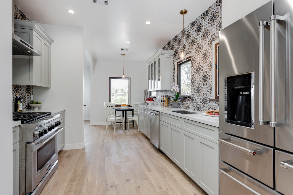 Photo of a mid-sized mediterranean galley eat-in kitchen in Los Angeles with recessed-panel cabinets, white cabinets, granite benchtops, multi-coloured splashback, no island, a single-bowl sink, terra-cotta splashback, stainless steel appliances, light hardwood floors and brown floor.