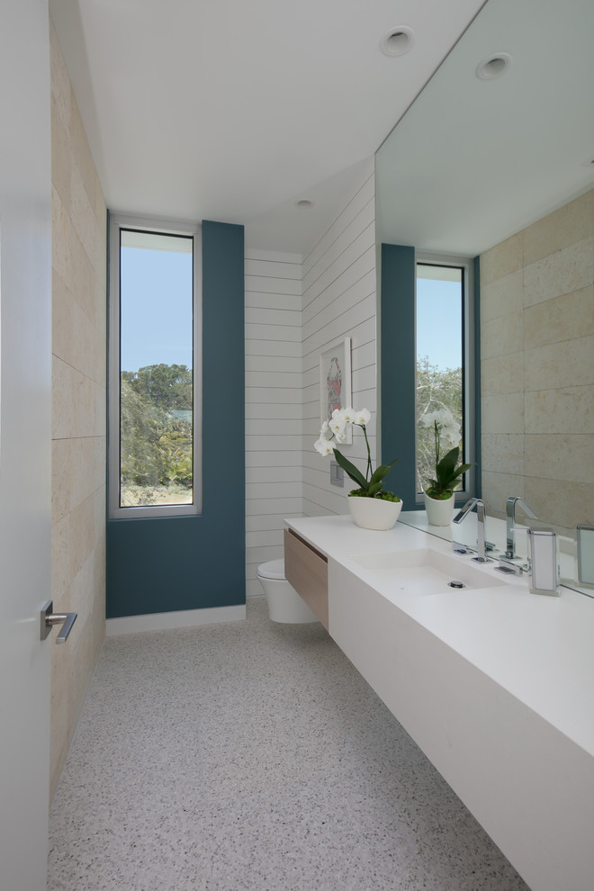 This is an example of a mid-sized beach style powder room in Tampa with flat-panel cabinets, light wood cabinets, a wall-mount toilet, limestone, multi-coloured walls, concrete floors, an integrated sink, solid surface benchtops, multi-coloured floor, white benchtops and beige tile.