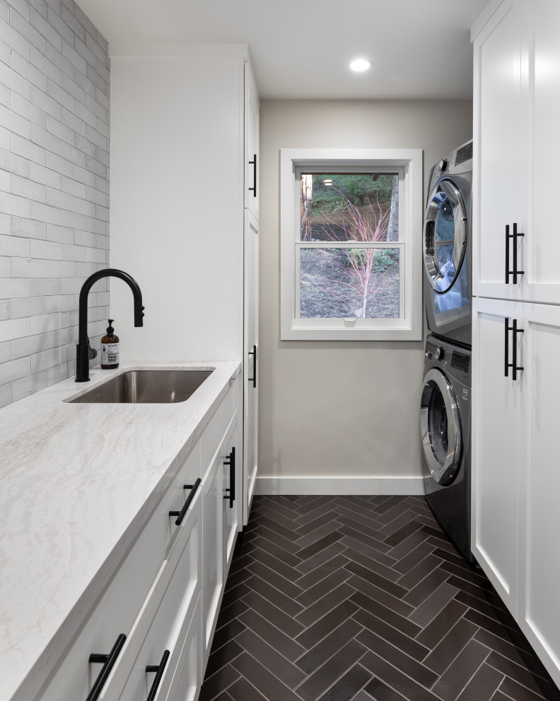 Inspiration for a mid-sized transitional galley dedicated laundry room in Orange County with an undermount sink, white cabinets, quartz benchtops, grey splashback, ceramic splashback, ceramic floors, a stacked washer and dryer, black floor, white benchtop, shaker cabinets and grey walls.