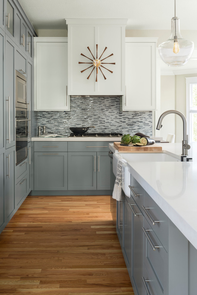 This is an example of a transitional kitchen in Denver with a farmhouse sink, shaker cabinets, grey cabinets, multi-coloured splashback, matchstick tile splashback, stainless steel appliances, with island, white benchtop and light hardwood floors.