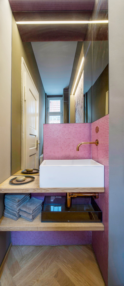 Photo of a small contemporary powder room in Stuttgart with open cabinets, pink tile, mosaic tile, pink walls, medium hardwood floors, a vessel sink, wood benchtops and beige benchtops.