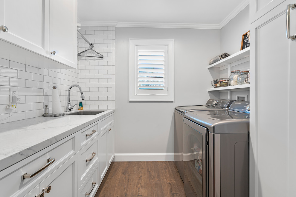 This is an example of a mid-sized transitional galley dedicated laundry room in San Francisco with an undermount sink, recessed-panel cabinets, white cabinets, marble benchtops, grey walls, dark hardwood floors, a side-by-side washer and dryer, brown floor and white benchtop.