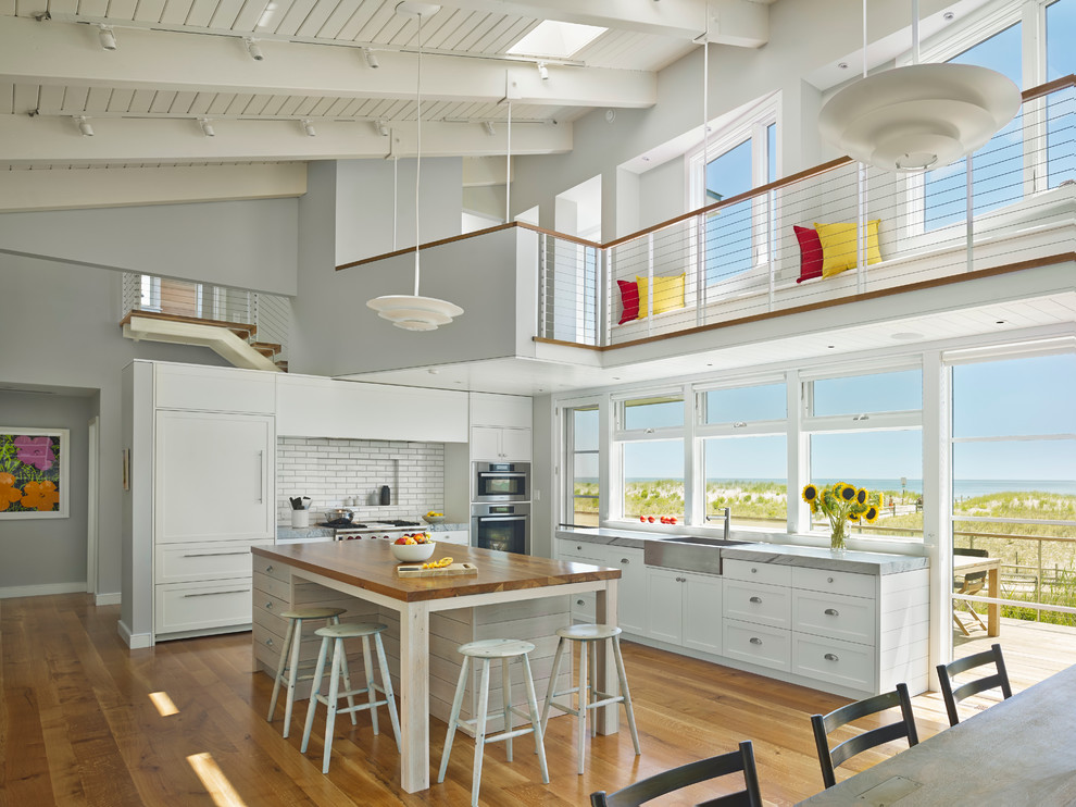 Inspiration for a mid-sized beach style l-shaped open plan kitchen in Philadelphia with a farmhouse sink, shaker cabinets, white cabinets, white splashback, subway tile splashback, medium hardwood floors, with island, marble benchtops, stainless steel appliances, brown floor and grey benchtop.