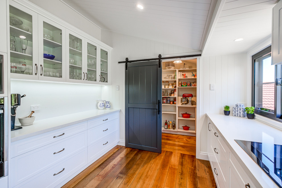 This is an example of an expansive contemporary galley kitchen/diner in Brisbane with a double-bowl sink, shaker cabinets, white cabinets, engineered stone countertops, white splashback, engineered quartz splashback, stainless steel appliances, light hardwood flooring, an island, brown floors, white worktops and a wood ceiling.
