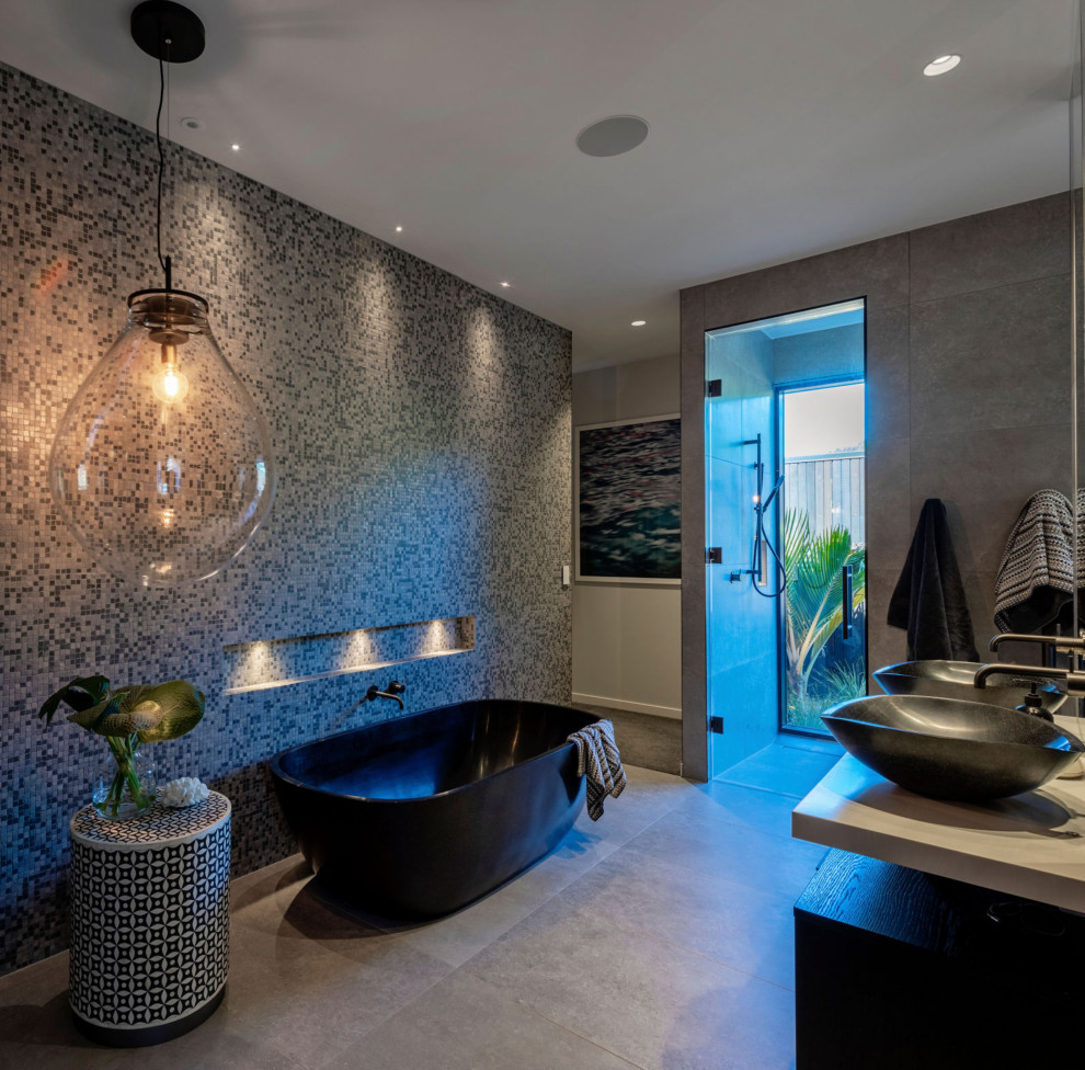 This is an example of a large contemporary master bathroom in Auckland with flat-panel cabinets, dark wood cabinets, a freestanding tub, an open shower, a two-piece toilet, multi-coloured tile, pebble tile, white walls, ceramic floors, a pedestal sink, engineered quartz benchtops, beige floor, an open shower, white benchtops, a niche, a double vanity, a floating vanity and wallpaper.