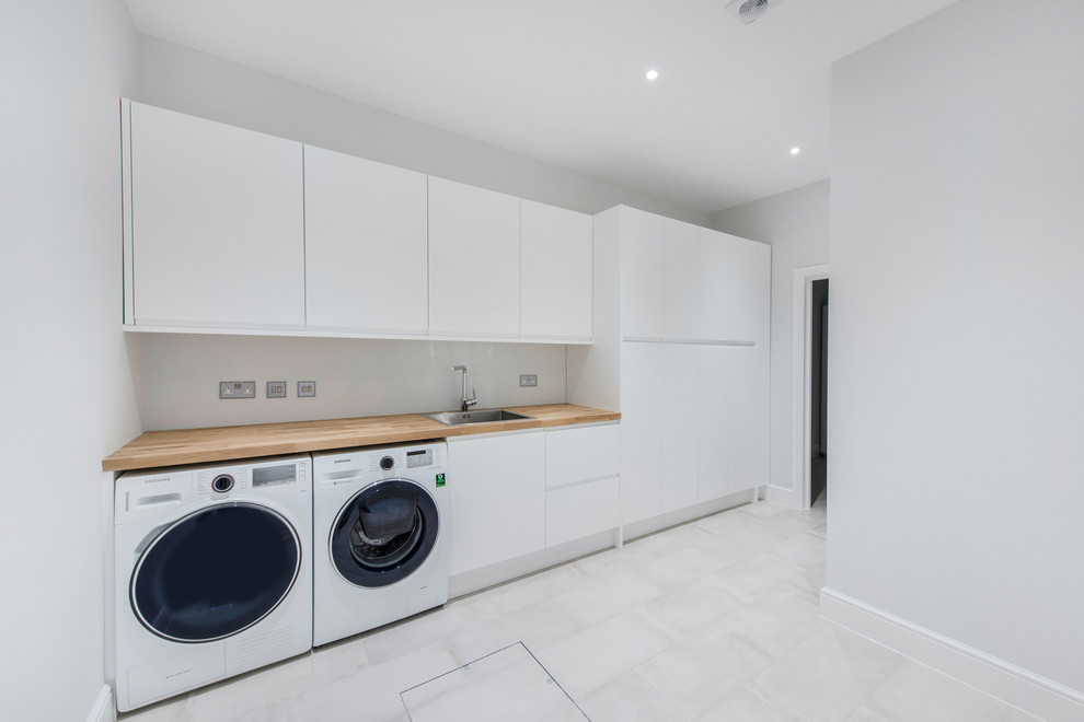 Inspiration for a large contemporary single-wall utility room in London with flat-panel cabinets, white cabinets, wood benchtops, white walls, a side-by-side washer and dryer, an integrated sink and ceramic floors.