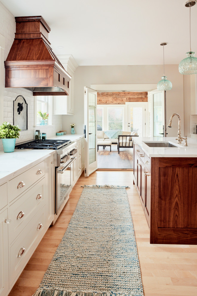 This is an example of a large rural kitchen/diner in Portland Maine with a single-bowl sink, shaker cabinets, white cabinets, engineered stone countertops, white splashback, ceramic splashback, stainless steel appliances, light hardwood flooring, an island and white worktops.