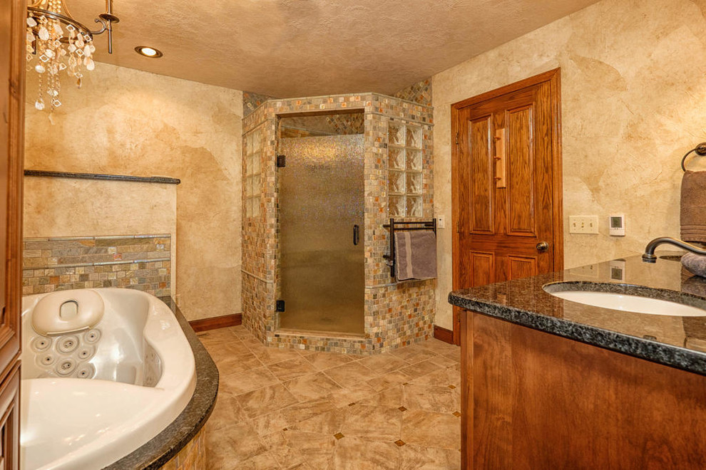 This is an example of a mid-sized country master bathroom in Minneapolis with an undermount sink, raised-panel cabinets, medium wood cabinets, granite benchtops, a drop-in tub, a corner shower, a one-piece toilet, multi-coloured tile, stone tile, beige walls and porcelain floors.
