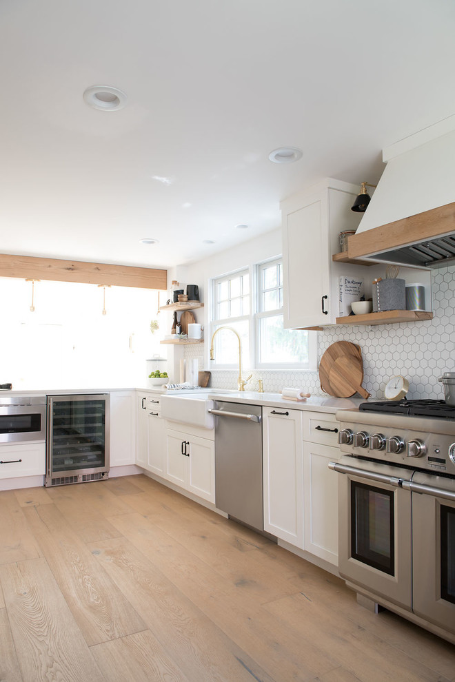 This is an example of a large country u-shaped eat-in kitchen in Boston with shaker cabinets, white cabinets, quartz benchtops, white splashback, mosaic tile splashback, stainless steel appliances, light hardwood floors, a peninsula, brown floor and white benchtop.