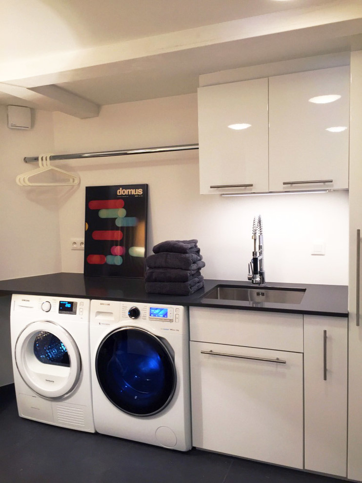 Mid-sized modern dedicated laundry room in Other with a single-bowl sink, white cabinets, quartzite benchtops, white walls, porcelain floors, a side-by-side washer and dryer, black benchtop and flat-panel cabinets.