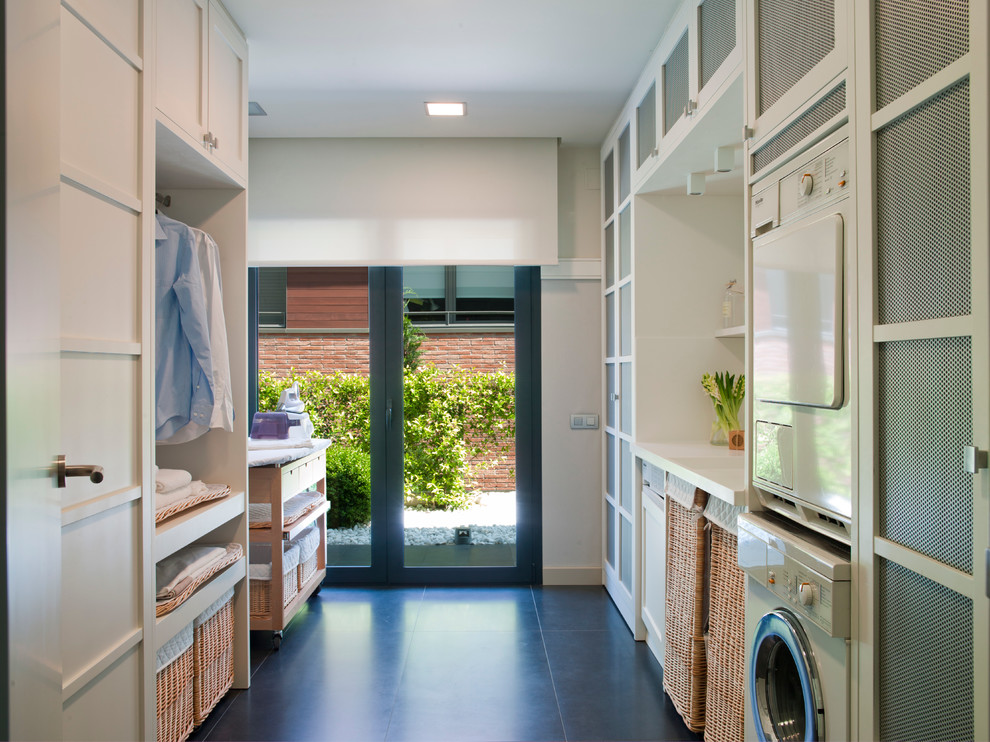 Design ideas for a mid-sized transitional galley dedicated laundry room in Barcelona with recessed-panel cabinets, white cabinets, solid surface benchtops, white walls, ceramic floors and a stacked washer and dryer.