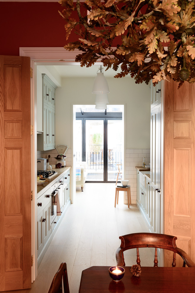 Design ideas for a mid-sized traditional galley separate kitchen in London with a double-bowl sink, beaded inset cabinets, blue cabinets, wood benchtops, white splashback, subway tile splashback, stainless steel appliances, light hardwood floors and no island.