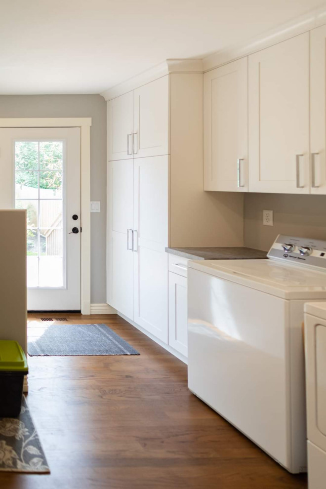This is an example of a large transitional l-shaped utility room in Seattle with a drop-in sink, shaker cabinets, white cabinets, laminate benchtops, white splashback, porcelain splashback, white walls, dark hardwood floors, a side-by-side washer and dryer, brown floor and grey benchtop.