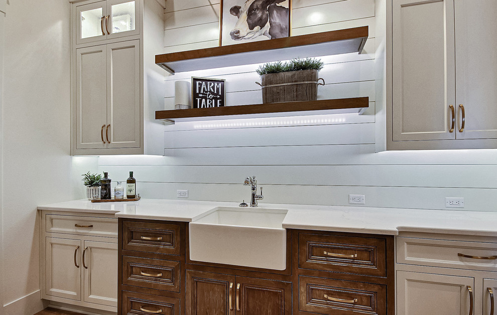 Photo of an expansive country u-shaped kitchen pantry in Portland with a farmhouse sink, recessed-panel cabinets, white cabinets, quartz benchtops, white splashback, ceramic splashback, stainless steel appliances, light hardwood floors, with island, brown floor and white benchtop.