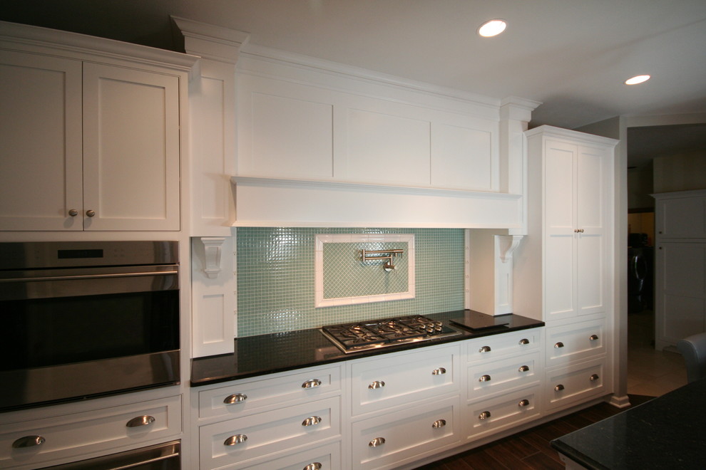 Mid-sized transitional u-shaped porcelain tile and brown floor enclosed kitchen photo with a farmhouse sink, shaker cabinets, white cabinets, quartzite countertops, green backsplash, glass tile backsplash, white appliances, an island and black countertops