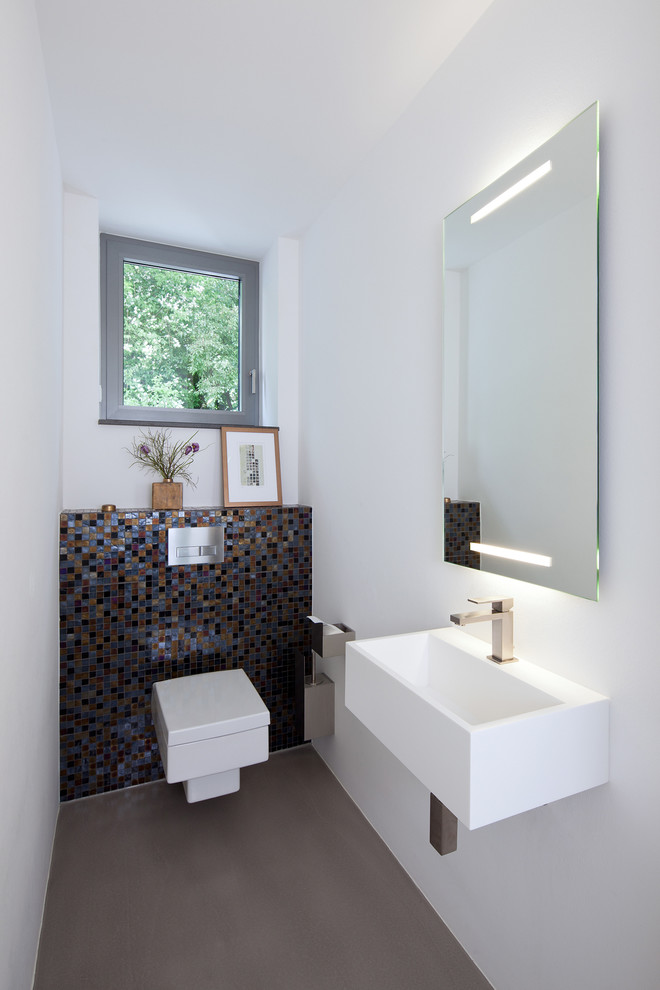 Inspiration for a mid-sized contemporary powder room in Essen with a wall-mount sink, multi-coloured tile, mosaic tile, white walls and a wall-mount toilet.