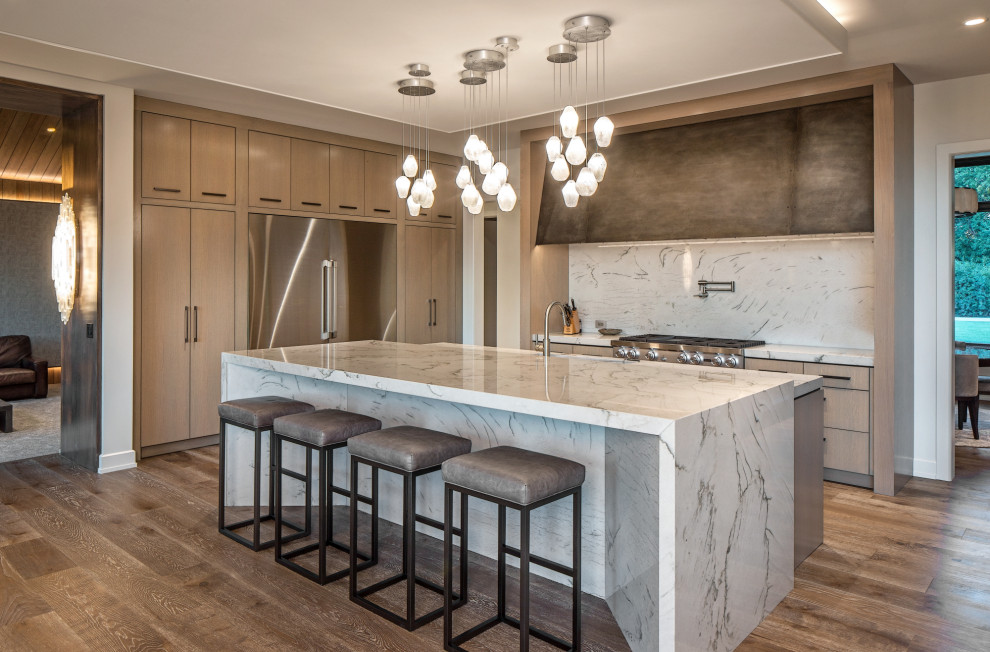 Inspiration for a country l-shaped kitchen in Omaha with flat-panel cabinets, light wood cabinets, white splashback, stone slab splashback, stainless steel appliances, medium hardwood floors, with island, brown floor and white benchtop.