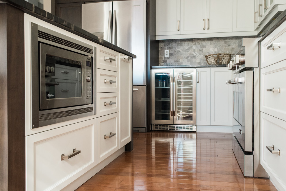 Inspiration for a transitional medium tone wood floor and brown floor kitchen remodel in Toronto with an undermount sink, recessed-panel cabinets, white cabinets, granite countertops, gray backsplash, travertine backsplash, stainless steel appliances and an island
