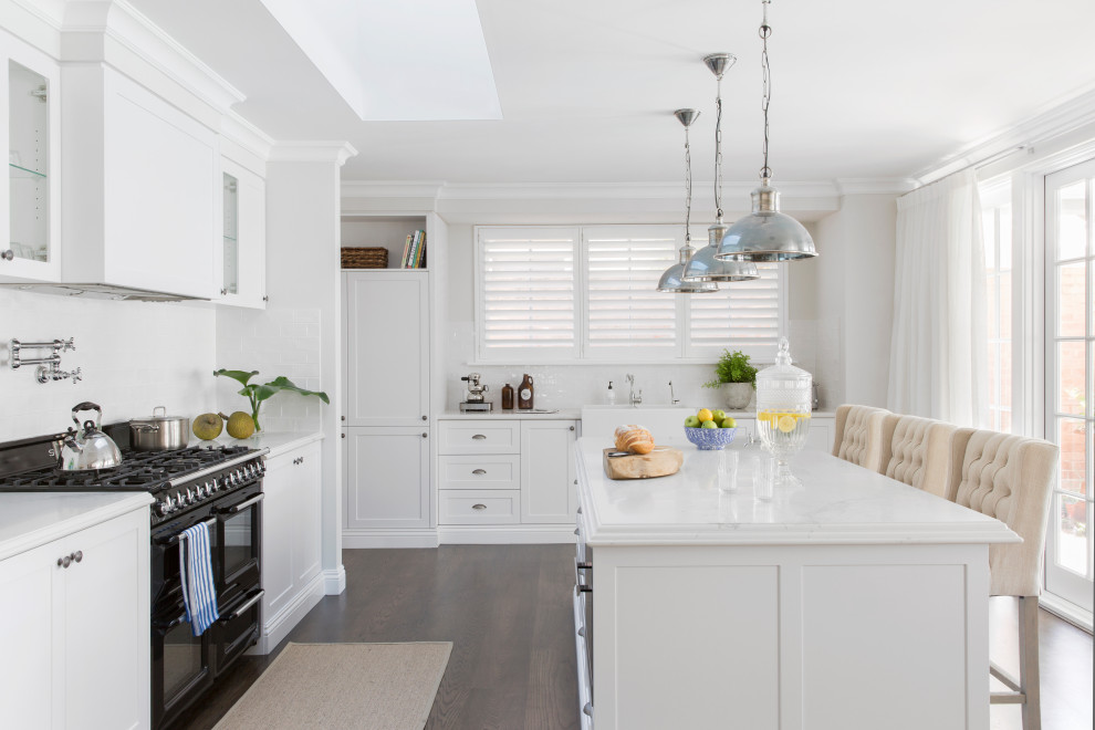 Inspiration for a transitional l-shaped kitchen in Perth with a farmhouse sink, shaker cabinets, white cabinets, white splashback, mosaic tile splashback, black appliances, dark hardwood floors, with island and white benchtop.