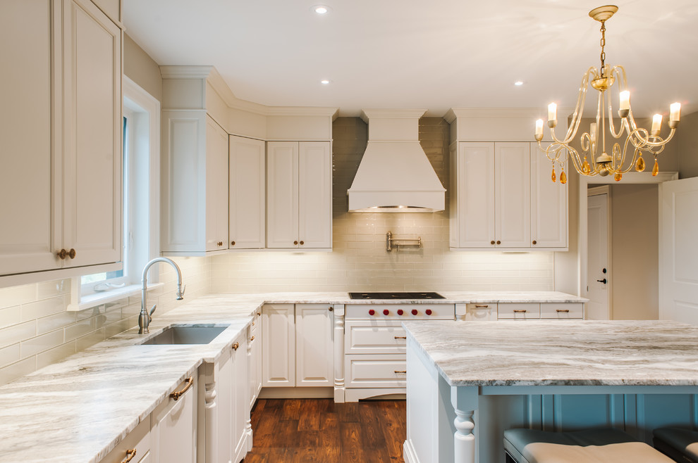 This is an example of a large traditional l-shaped eat-in kitchen in Toronto with an undermount sink, raised-panel cabinets, white cabinets, quartzite benchtops, white splashback, glass tile splashback, medium hardwood floors, with island, brown floor and beige benchtop.