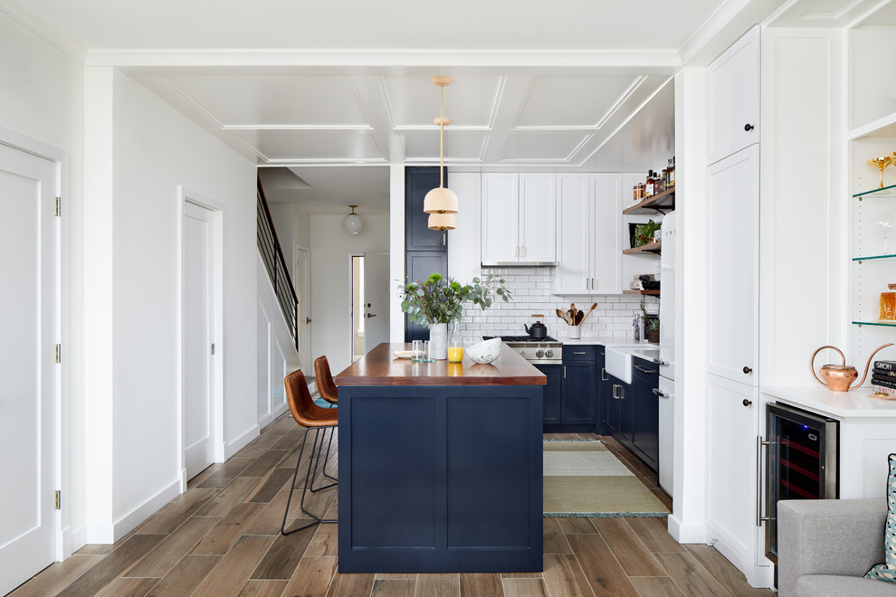 Inspiration for a small eclectic l-shaped open plan kitchen in DC Metro with a farmhouse sink, shaker cabinets, blue cabinets, wood benchtops, white splashback, ceramic splashback, stainless steel appliances, porcelain floors, with island and brown floor.