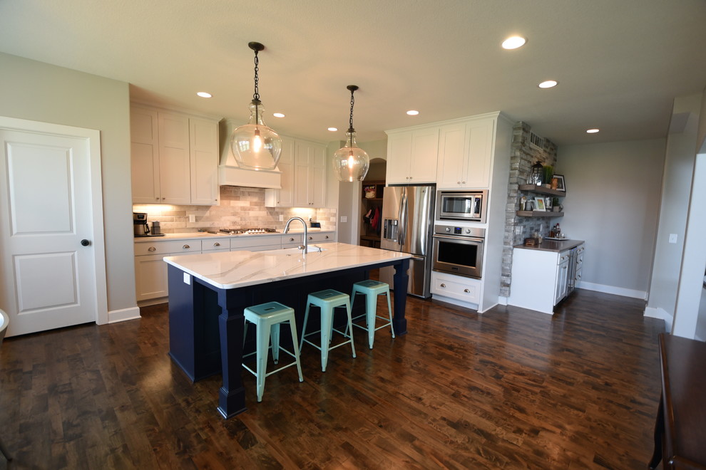 This is an example of a transitional l-shaped eat-in kitchen in Kansas City with a farmhouse sink, shaker cabinets, white cabinets, quartz benchtops, grey splashback, ceramic splashback, stainless steel appliances, dark hardwood floors, with island, brown floor and white benchtop.