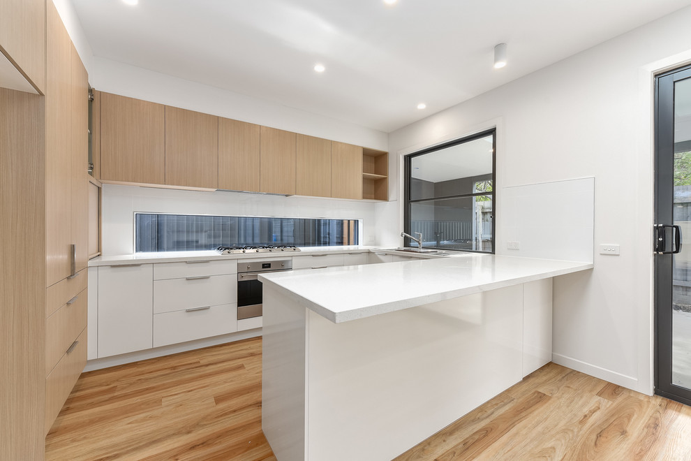 Design ideas for a mid-sized modern u-shaped eat-in kitchen in Melbourne with a double-bowl sink, light wood cabinets, white splashback, porcelain splashback, stainless steel appliances, vinyl floors, no island, brown floor and white benchtop.