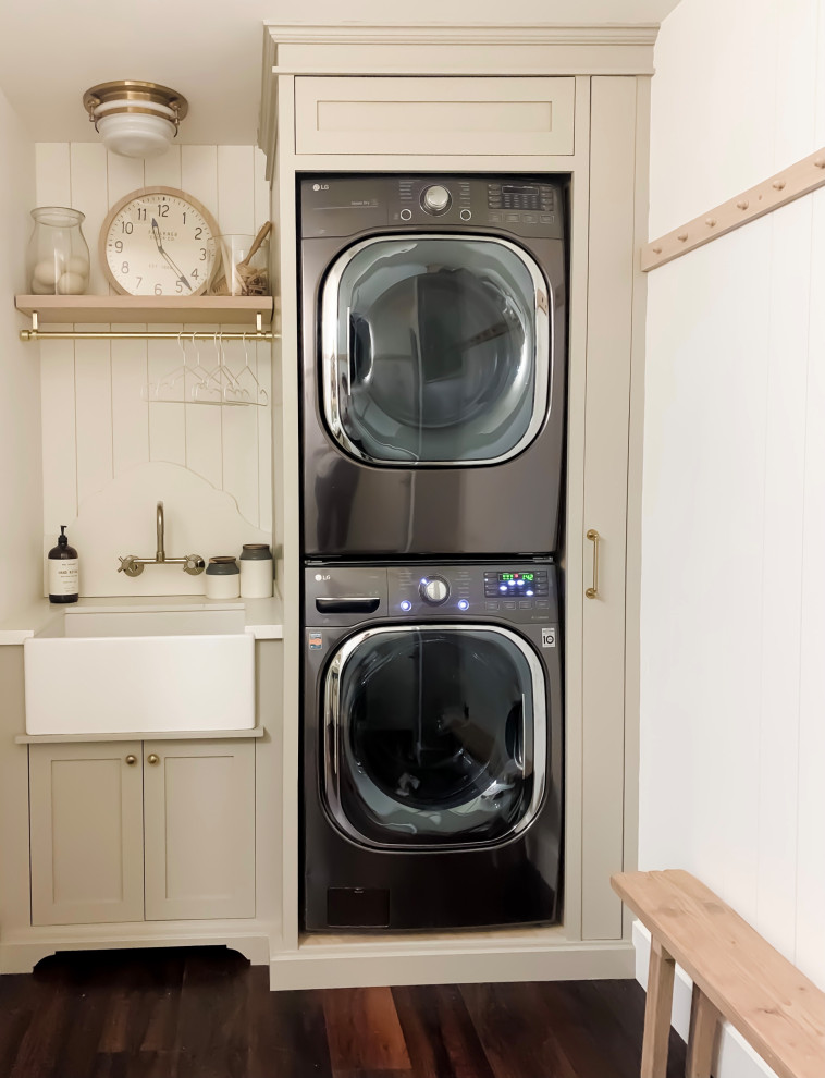 Mid-sized traditional single-wall dedicated laundry room with a farmhouse sink, shaker cabinets, beige cabinets, quartz benchtops, timber splashback, white walls, vinyl floors, a stacked washer and dryer, brown floor, white benchtop and planked wall panelling.