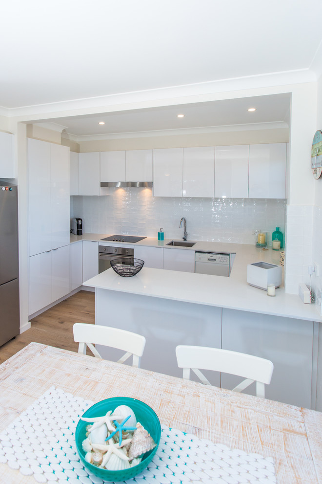 Inspiration for a small beach style u-shaped eat-in kitchen in Sydney with a drop-in sink, flat-panel cabinets, white cabinets, quartz benchtops, white splashback, subway tile splashback, stainless steel appliances, light hardwood floors and no island.