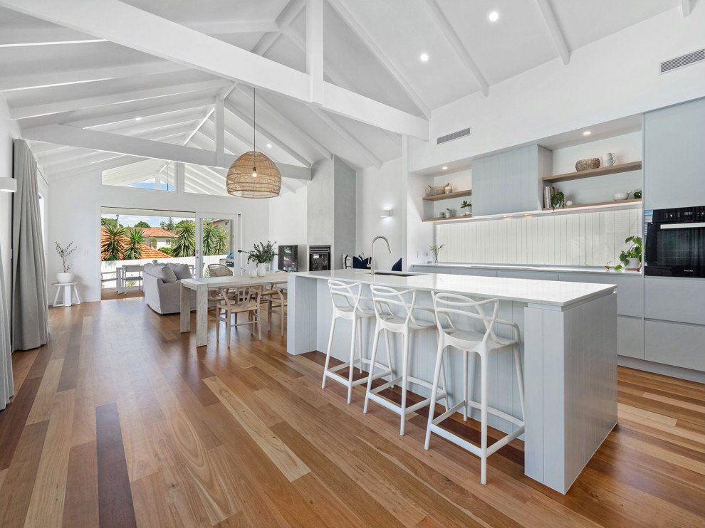 Mid-sized beach style galley open plan kitchen in Brisbane with an undermount sink, flat-panel cabinets, window splashback, black appliances, with island, brown floor, exposed beam, vaulted, grey cabinets, medium hardwood floors and white benchtop.