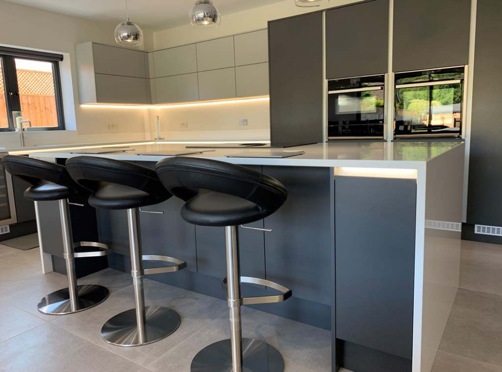 This is an example of a modern l-shaped open plan kitchen in Dorset with a drop-in sink, flat-panel cabinets, grey cabinets, quartzite benchtops, white splashback, engineered quartz splashback, black appliances, with island and white benchtop.