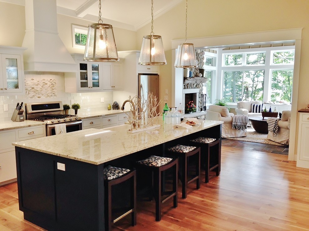 This is an example of a beach style single-wall kitchen in Grand Rapids with a farmhouse sink, shaker cabinets, white cabinets, granite benchtops, white splashback, glass tile splashback, stainless steel appliances, medium hardwood floors and with island.