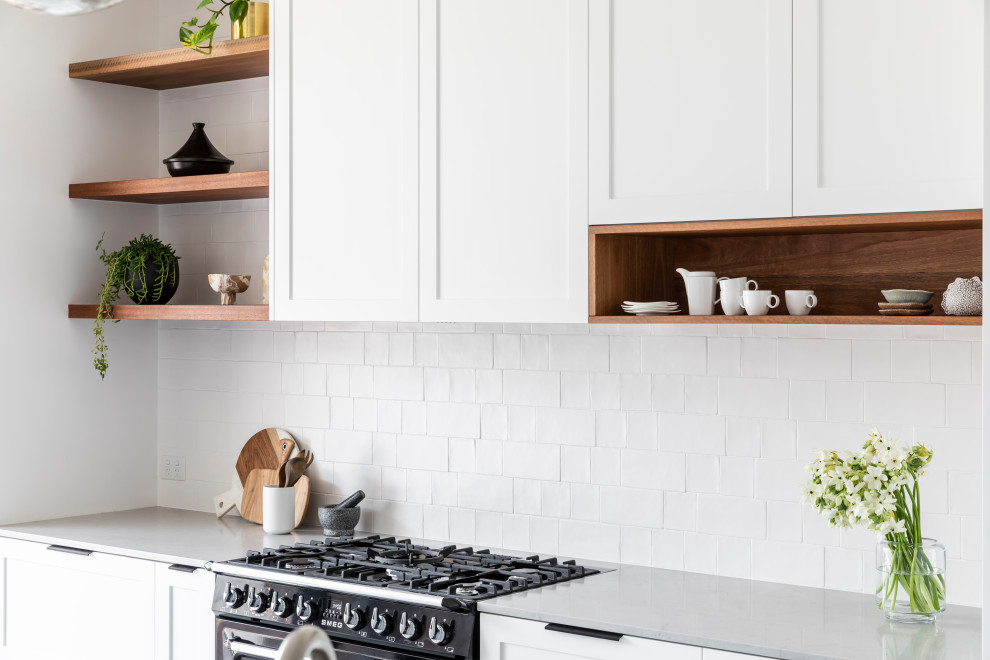 This is an example of a mid-sized transitional single-wall open plan kitchen in Sydney with an undermount sink, shaker cabinets, white cabinets, quartz benchtops, white splashback, ceramic splashback, black appliances, concrete floors, with island, grey floor and grey benchtop.