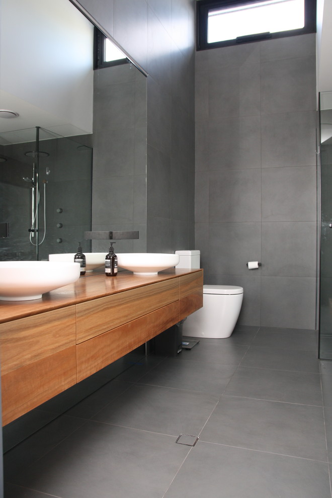 Inspiration for a large contemporary master bathroom in Adelaide with furniture-like cabinets, medium wood cabinets, a freestanding tub, an open shower, a one-piece toilet, gray tile, cement tile, grey walls, cement tiles, a vessel sink, wood benchtops, grey floor and a hinged shower door.