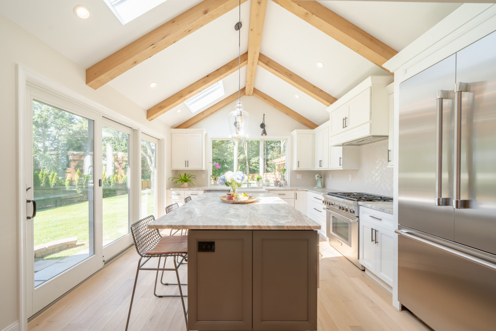 Eat-in kitchen - large transitional l-shaped light wood floor, brown floor and exposed beam eat-in kitchen idea with a farmhouse sink, shaker cabinets, white cabinets, white backsplash, stainless steel appliances, an island, multicolored countertops, marble countertops and ceramic backsplash