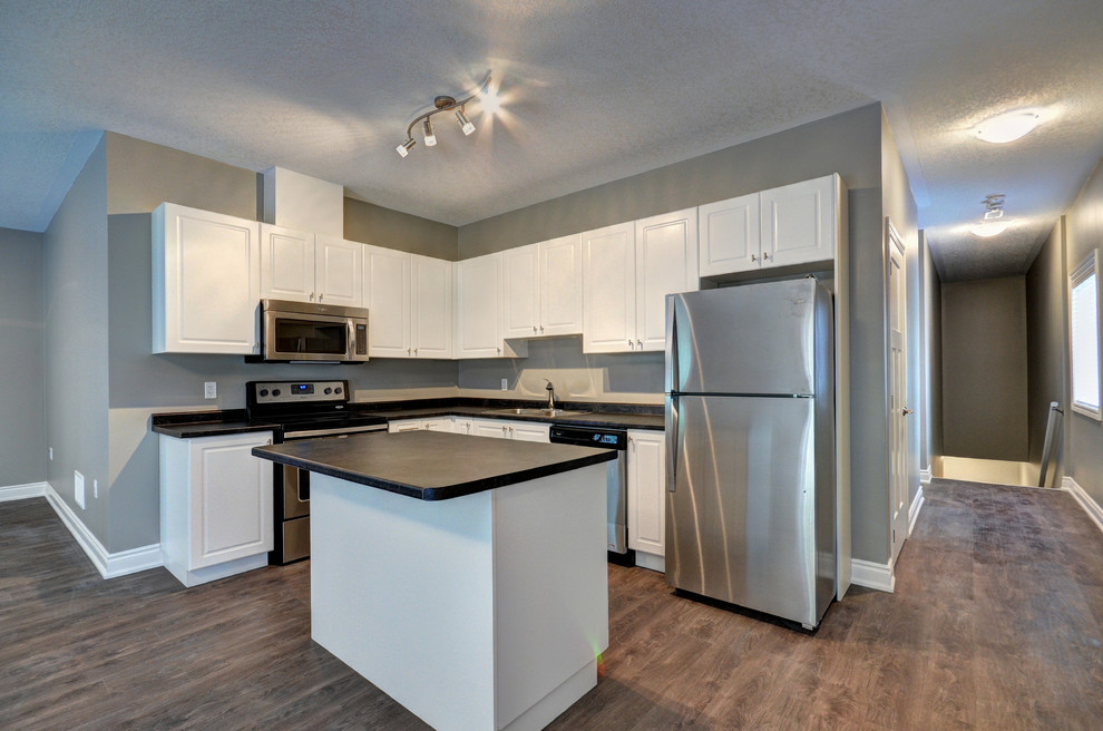 Medium sized modern l-shaped kitchen/diner with a built-in sink, shaker cabinets, white cabinets, stainless steel appliances, dark hardwood flooring and an island.